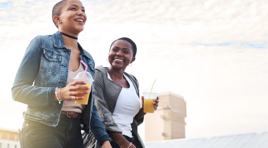 Friends, together and walking with blue sky and drinks for wellness, outdoor with happiness for freedom. Women, laugh and smile in neighborhood with discussion on equality, rights and opportunity.